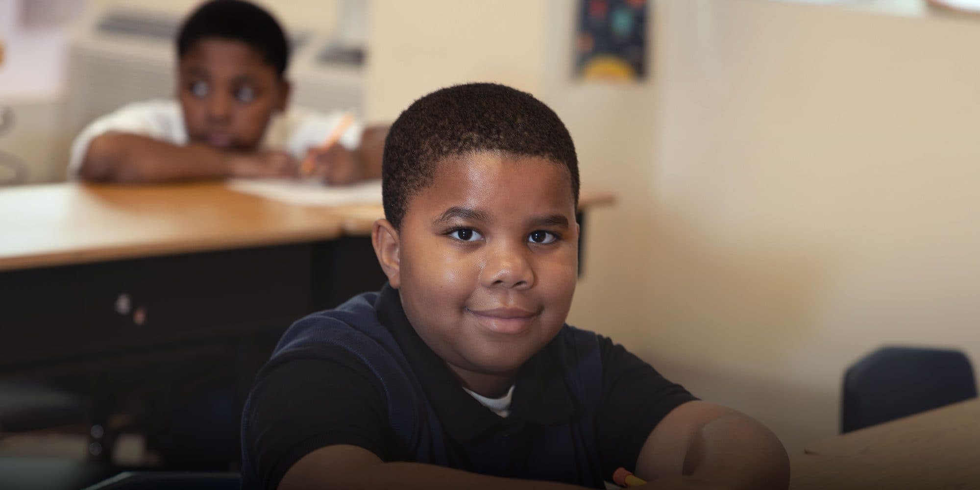 Smiling student in a navy polo sitting at a classroom desk.