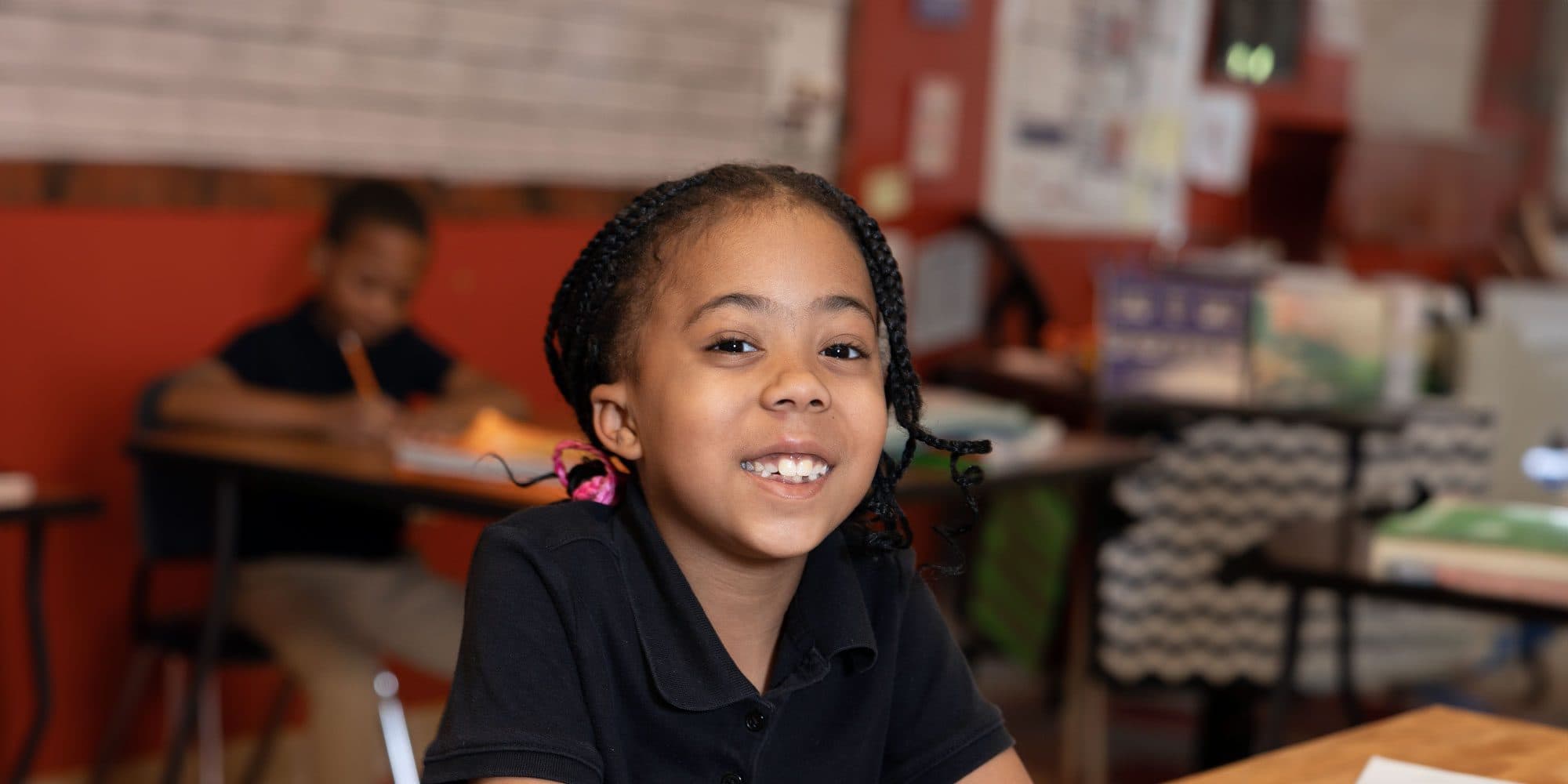 Smiling student with braided hair sitting at a desk in a classroom.