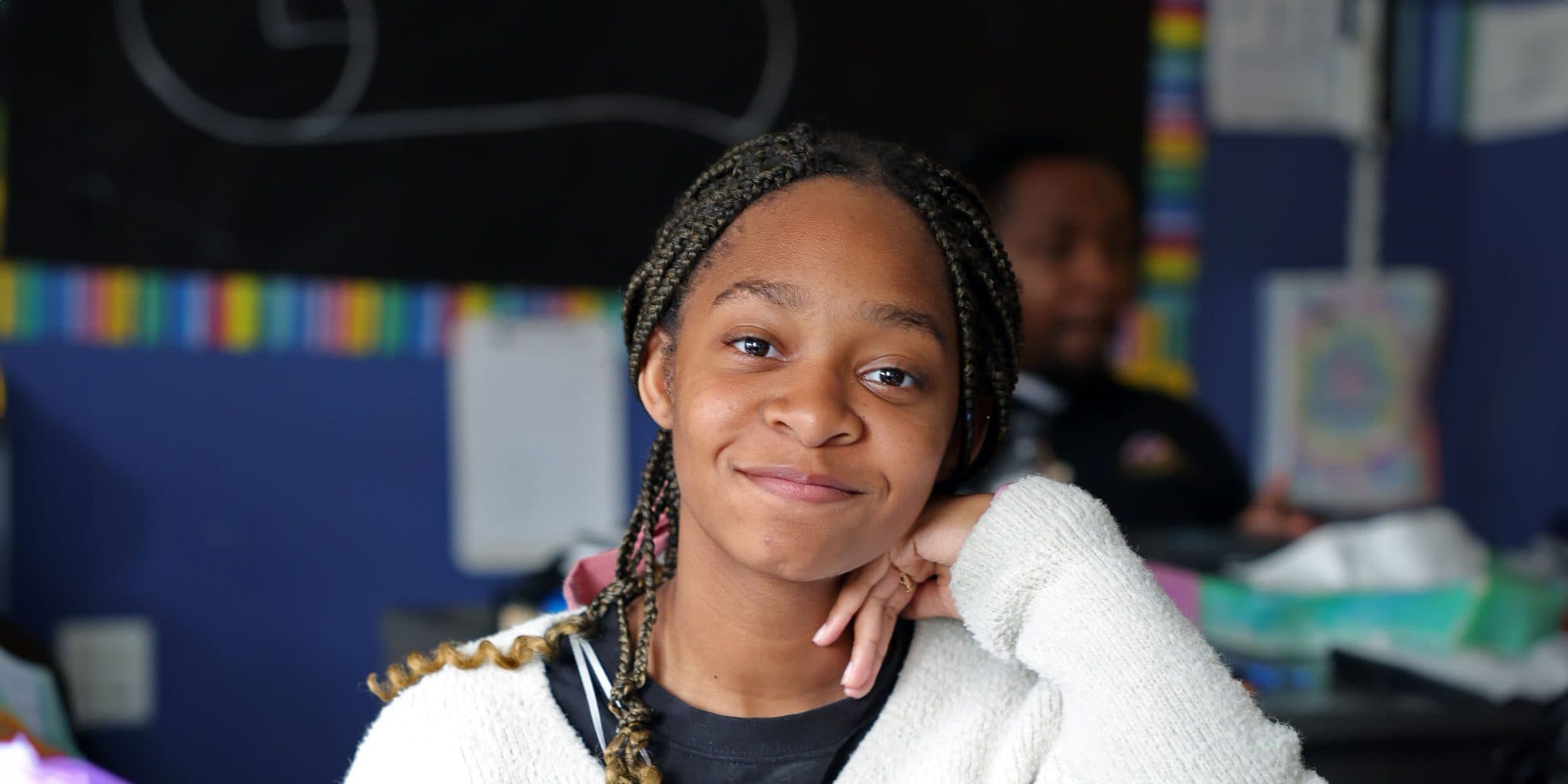 Smiling student with braided hair resting their chin on their hand in a classroom.